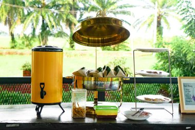 Açık büfe tezgahında sarı içecek dağıtıcısı, muz yaprağı sarılı Nasi Lemak, hamur işleri, ve tropikal palmiye ağaçları ve tarlaları..