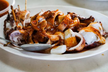 Meşhur bir Malezya deniz ürünleri yemeği olan ızgara doldurulmuş mürekkep balığının yakın çekimi bir paket servis konteynırında servis edildi. Baharat zengini, sulu ve tamamen kömürleşmiş..