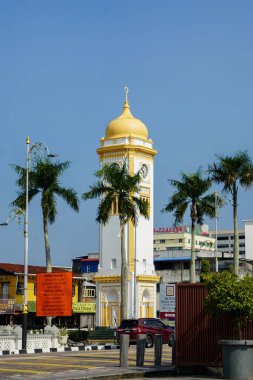 Kedah, Malezya, 5 Aralık 2025: Altın kubbeli beyaz ve sarı bir saat kulesi Alor Setar 'da palmiye ağaçları, dükkanlar ve oteller arasında duruyor, miras ve şehir yaşamını harmanlıyor..