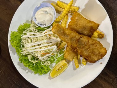 Haşlanmış altın sarısı balık filetosu taze lahana salatasıyla kıtır kıtır patates kızartması ve sıcak bir yemek ortamında beyaz bir tabakta limon dilimi servis edilir..