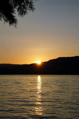 Sakin bir gölün üzerinde huzurlu bir günbatımı. Altından yansımalar, siluetli ağaç dalları ve ılık ve serin tonlarla dolu bir gökyüzünün altındaki uzak tepeler..
