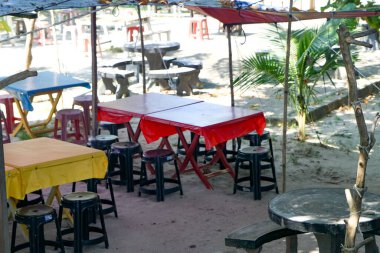 Kırmızı, sarı ve mavi masa örtüleriyle kaplı, siyah ve kırmızı taburelerle çevrili bir açık hava oturma alanı..