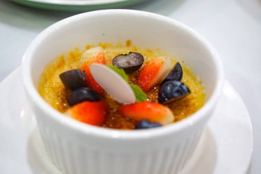 Çilek, yaban mersini, üzüm, nane ve karamelize şeker kabuğuyla kaplı beyaz bir ramekin içerisindeki zarif krema brulee, beyaz bir çay tabağında servis edilir..