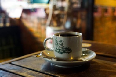 Çiçek tabağı ve kaşığın üzerinde koyu kahve olan seramik fincan, rahat kafeterya ortamında ahşap masa üzerine kurulmuş..