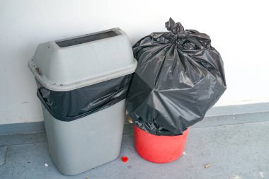 İki çöp bidonu yan yana gri bir zeminde beyaz bir duvara karşı, biri düzgün bir şekilde kullanılmış ve biri taşmış, atık yönetimi ve temizliği ön plana çıkarıyor..