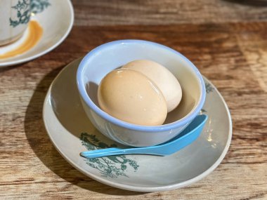 İki kahverengi yumurta, mavi kenarlı beyaz bir kasenin içine yerleştirilmiş çay tabağı ve kaşık, ahşap masadaki sade yemek düzeni..