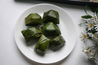 Beyaz bir tabakta muz yaprakları ve çiçek arkaplanıyla sunulan özgün Malezya tatlı kültürü sergilenmektedir..