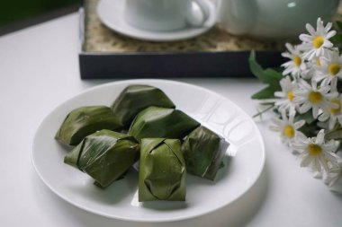 Beyaz bir tabakta muz yaprakları ve çiçek arkaplanıyla sunulan özgün Malezya tatlı kültürü sergilenmektedir..