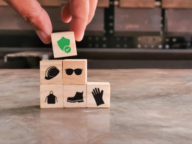 Bir el, çeşitli Kişisel Koruyucu Ekipman simgelerinin yer aldığı bir yığın tahta bloğun üzerinde kontrol işareti olan bir kalkan simgesini yerleştiriyor..