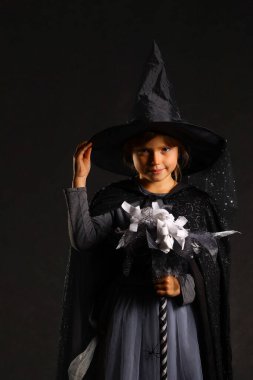 Cadı kostümlü gülümseyen küçük bir kız sivri uçlu bir şapka ve Cadılar Bayramı süpürgesi çocuklar için şirin bir fantezi kıyafeti tutuyor.