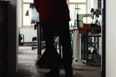 Eski atölyedeki adam dolu bir el arabasını masa ve büyük bir kutuyla karışık endüstriyel alanda kameraya doğru itiyor.