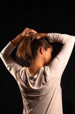 Saçlarını iki eliyle at kuyruğuna çeviren kadın, arkadan görülüyor, günlük kişisel bakım rutininin bir parçası, makyaj, kadınsı enerjinin sakin anı ve kişisel zamanı.