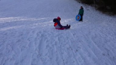 Genç bir kız, plastik bir kızakla karlı bir kış yamacında hızla kayıyor, çocukluk kış eğlencesi ve açık hava oyunu, büyük bir oğlan yokuş yukarı yürürken kamerayı geçiriyor.