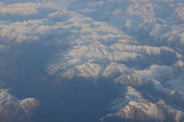 Karla kaplı dağ zirveleri, güneş ışığı ve gölgeli tepeler yüksek irtifa perspektifinden kışın engebeli dağlık araziyi vurguluyor.
