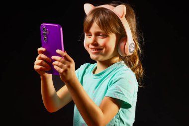 Çocuk elinde akıllı bir telefonla gülümsüyor ve karanlık, modern bir ortamda oyuncu kategorisinde kulaklık takıyor, çocukluk neşesini, dijital etkileşimi ve çağdaş çocukların teknolojisini vurguluyor.