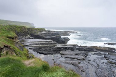 İrlanda 'daki Mohor kayalıklarının panoramik manzarası. Dalgalı Atlantik Okyanusu ve kayalık bir kıyı şeridi.