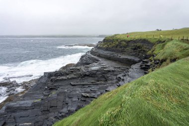 İrlanda 'daki Mohor uçurumlarının panoramik görüntüsü. Dalgalı bir Atlantik okyanusu ve bazı yürüyen insanlarla dolu kayalık bir sahil şeridi..