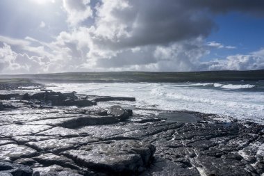 İrlanda 'daki Doolin İskelesi. Mavi gökyüzünün altında bulutlu kayalık bir manzara. Arka planda Atlantik Okyanusu.