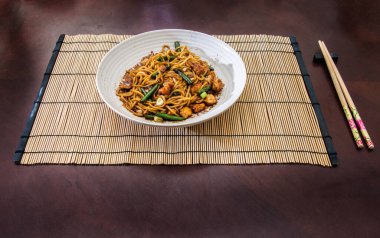 Bambu hasırla servis edilen bir kase tavuk kızartmalı erişte. İçinde renkli sebzeler ve Asya 'dan esinlenilmiş bir sunum var..