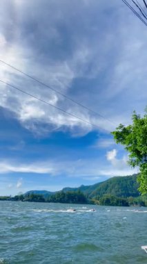 Sarangan Gölü 'ndeki dağlık manzaranın güzel manzarası. Parlak mavi gökyüzünün altında sık orman zirveleri var. Uçsuz bucaksız, dalgalı bir göl ön planda, suda dalgalar, doğanın güzelliği ve açık hava eğlencesi yaratan küçük teknelerin yer aldığı yerde..