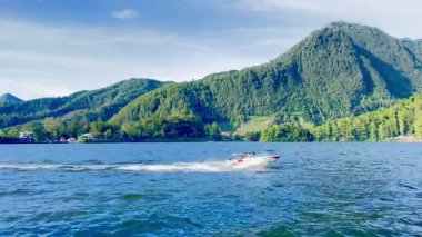 Sarangan Gölü 'ndeki dağlık manzaranın güzel manzarası. Parlak mavi gökyüzünün altında sık orman zirveleri var. Uçsuz bucaksız, dalgalı bir göl ön planda, suda dalgalar oluşturan küçük tekneler barındırıyor. Çevredeki tepeler yemyeşil ve yemyeşil
