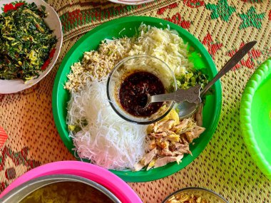 Yeşil plastik leğen, Asya mutfağıyla ilişkili çeşitli malzemelerle birlikte. Parçalanmış lahana, fasulye filizi, ince vermicelli, doğranmış tavuk ve cam kasede acı biberli ya da soya soslu kalın sos var. Tabağa bir kaşık konur.