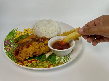 Patates kızartmasını küçük bir kase sosa batıran birinin yakın çekimi. Arka plandaki yuvarlak tabakta beyaz pirinç, mısır ve yanında patates kızartması var. Tabakta yeşil bir yaprak tasarımı var. Elleri bir yetişkinin ellerine benziyordu.,