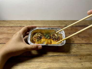 Bir insan, mentai sosu ve otlarla kaplı bir parça Dim Sum 'u tutmak için çubuk kullanır. Dim Sum, ahşap bir masada küçük bir folyo kabının içinde. Odak, kaldırılan parçanın dokusunu ve malzemelerini vurguluyor..