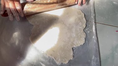 Eller un kaplı bir yüzeyde tahta bir oklava ile hamuru yuvarlıyor. Kişi sıkıca kavrar, parmakları saplarının üzerine yayılır, un ile toz alır. Hamur dikdörtgen ve pürüzsüz, şeffaf bir yüzey üzerinde düz, fayanslı bir tezgah üzerinde