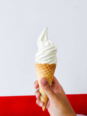 Bir el, waffle külahında kıvrılmış vanilyalı dondurma tutuyor. Koninin klasik elmas deseni ve dondurmanın da kremalı beyazı var. Arka plan beyaz bir üst bölüm ve kırmızı bir alt bölümden oluşur.,
