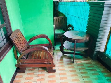 Yeşil boyalı duvarları ve fayanslı zemini olan küçük bir açık hava alanının köşesi. İçinde ahşap mobilyalar var: iki sandalye ve iki katlı bir yuvarlak masa. Bir turkuaz gizlilik ekranı görünür