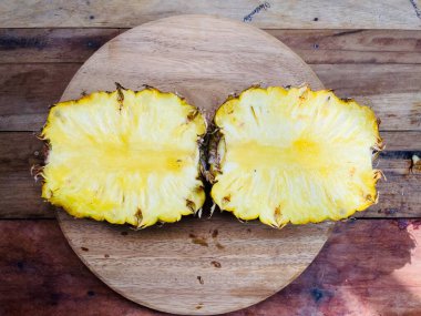 Ortadan ikiye bölünmüş bir ananas sulu, sarı etini yuvarlak bir tahta kesme tahtasının üzerinde sergiler. Ananasın pürüzlü dış kabuğu kısmen çıkarılmış ve lifli iç kısmı ortaya çıkmış. Meyvenin dokusu lifli ve nemlidir.,