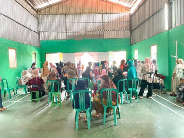 Kadınlar ve çocuklar, Endonezya 'da yerel bir sağlık hizmeti olan yeşil duvarlı bir sağlık merkezinde toplanıyorlar. Bazı kadınlar başörtüsü takar. Sağlık çalışanları bir masada ziyaretçilere yardım ediyor. Bir kadın bir ölçü aletinin yanında durur.