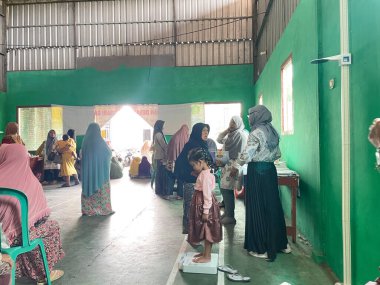 Endonezya 'da bir pozyandu içinde, renkli başörtüleri ve elbiseli kadınlar toplumsal sağlık faaliyetlerine katılıyorlar. Bir kız tartı tartısında duruyor, sağlık kontrolünden geçmesi muhtemel. Sağdaki masa, sağlık personeli için bir çalışma masası görevi görüyor..