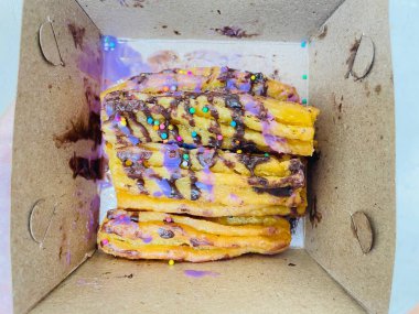 Birkaç churro açık bir karton kutunun içine yerleştiriliyor. Çikolata ve mor krema serpiştirilmiş renkli yuvarlak serpiştirilmiş. Karton kutu sade, görünür katlama çizgileri ve sekmeleri var..