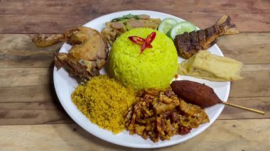 Beyaz tabakta, muhtemelen zerdeçal katılmış kırmızı biber dilimleriyle süslenmiş canlı sarı pirinç yığını bulunur. Pirincin etrafında çeşitli Endonezya yemekleri var, bir el satay koyuyor.