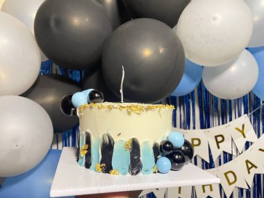 Yuvarlak bir doğum günü pastası, beyaz kremalı, siyah ve mavi soyut desenli ve altın yapraklı. Üzerinde bir mum ve mavi ve siyah top şeklinde dekorasyon koleksiyonu var. Arkaplanda siyah, beyaz ve mavi balonlar yer alıyor.