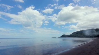 Sakin Mavi Denizi, Volkanik Karanlık Kumu ve Parlak Bulutlu Gökyüzü Altındaki Tepeleri olan Güzel Tropikal Plaj.