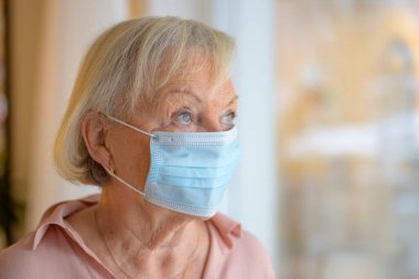 Coronavirus salgını sırasında tek kullanımlık cerrahi maske takan yaşlı bir kadın pencereden dikkatlice dışarı bakıyor.