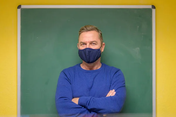 Confident serious male teacher with folded arms standing in front of a clean chalkboard in the classroom looking at camera