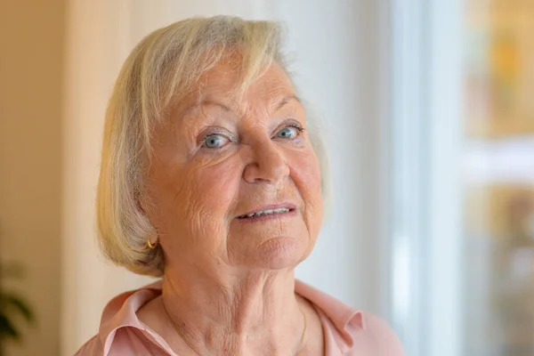 Quizzical senior lady glancing sideways at the camera as she stands near a large window at home in a close up head shot