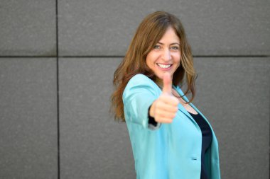 Motive olmuş, şevkli, orta yaşlı bir kadın. Telif boşluğu olan gri fayanslı duvarın üzerinde başarı dolu bir gülümsemeyle baş parmak işareti yapıyor.