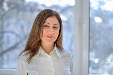 Beyaz bluzlu, çekici, orta yaşlı bir kadının doğal portresi. Pencerenin önünde durmuş, neşeli bir gülümsemeyle kameraya bakıyor.