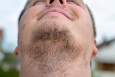 Unshaven young man with a wispy beard in close up on his throat and facial hair outdoors against grey sky clipart