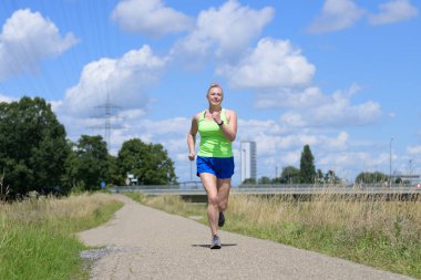 Sağlıklı, tombul, orta yaşlı bir kadın aktif bir yaşam tarzında, güneşli bir yaz gününde kırsal bir patika boyunca koşuyor ya da koşuyor.