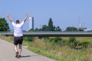 Son sınıf öğrencisi kameradan uzaklaşıp güneşli bir yaz gününde bir kanalın yanındaki patika boyunca V işareti yapıyor.