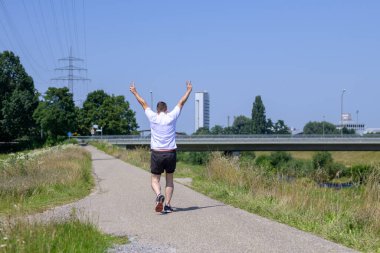 Kıdemli bir adam, güneşli bir yaz gününde, açık hava yaşam tarzı kavramında, bir patika boyunca V işareti yaparak kameradan uzaklaşıyor.