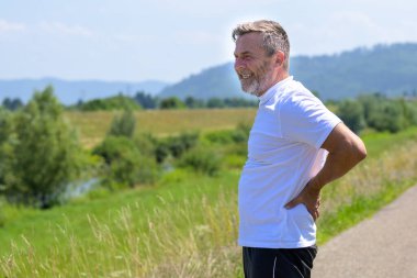 Orta yaşlı bir adam, güneşli bir yaz gününde kırsal bir yolda sağlık ve zindelik kavramında koşarken belini tutarken acı içinde kıvranıyor.