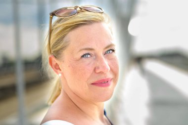 Gülümseyen, orta yaşlı, sarışın, güneş gözlüklü, açık havada, önemli bir portrede.