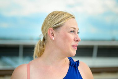 Mavi gözlü, sarışın, orta yaşlı bir kadın portresi. Yazın güneşli havada poz verirken yan yan bakıyor. Üstü başı mavi.